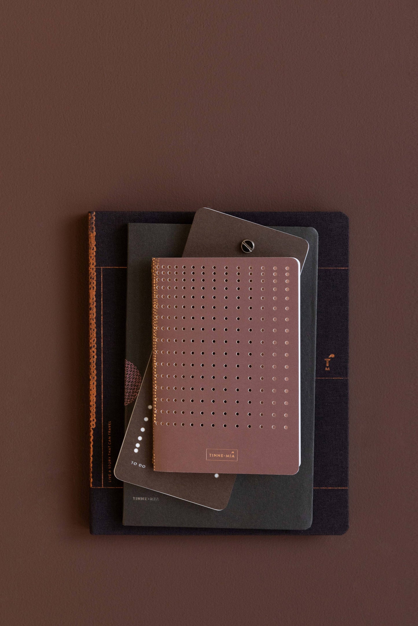 Ein Stapel von drei Tinne Mia - Bullet Journal A6-Notizbüchern, darunter eines mit burgunderrotem Rastereinband und eleganten Metallic-Akzenten, ist auf einer braunen Oberfläche angeordnet.