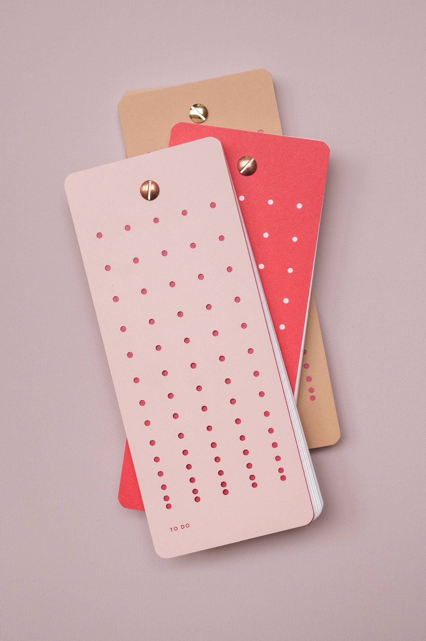 Drei Tinne Mia "Aufgabenliste - Nougat" Notizblöcke in Rosa, Rot und Hellbraun sind gestapelt und mit einer goldenen Klammer versehen. Sie sind aus FSC-zertifiziertem Papier hergestellt und haben perforierte, gepunktete Reihen. Der Hintergrund ist hell lila. Marke: Tinne Mia.