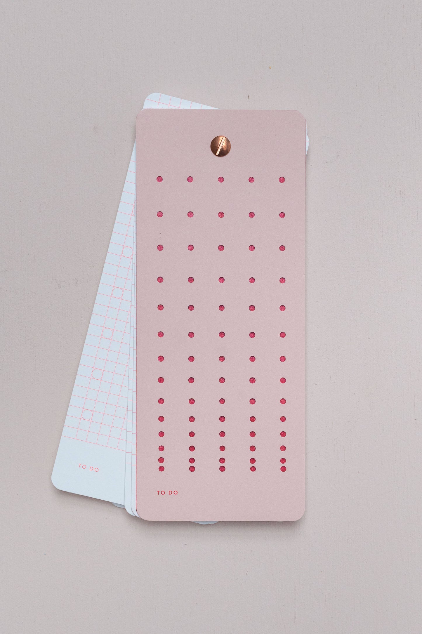 Die Tinne Mia - Aufgabenliste - Mellow Rose von Tinne Mia zeigt eine rosa Aufgabenliste mit roten Punkten auf einer weißen Gitterkarte, die von einem goldenen Verschluss gehalten wird. Hergestellt aus FSC-zertifiziertem Papier für einen umweltfreundlichen Touch.