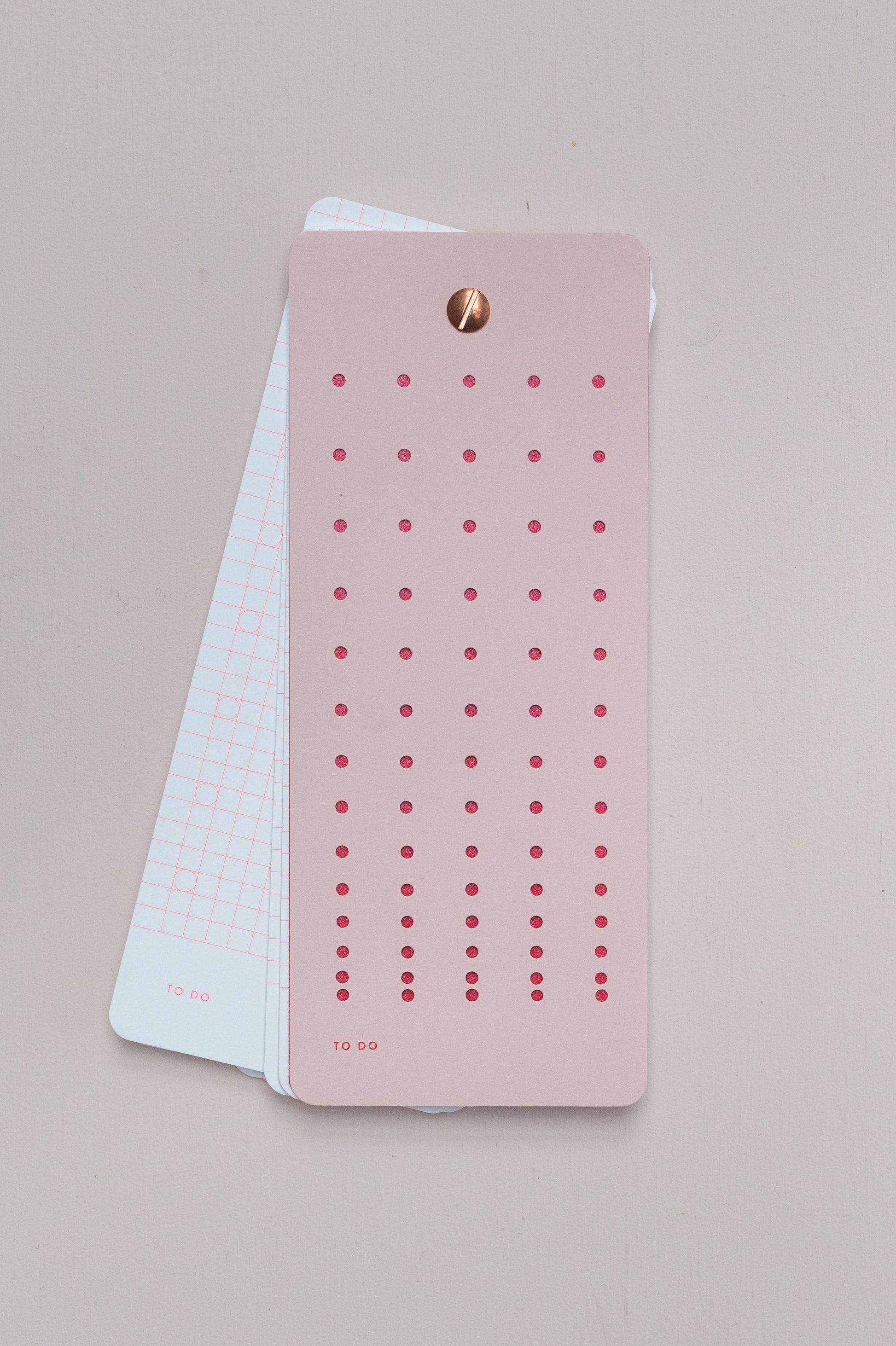 Die Tinne Mia - Aufgabenliste - Mellow Rose von Tinne Mia zeigt eine rosa Aufgabenliste mit roten Punkten auf einer weißen Gitterkarte, die von einem goldenen Verschluss gehalten wird. Hergestellt aus FSC-zertifiziertem Papier für einen umweltfreundlichen Touch.