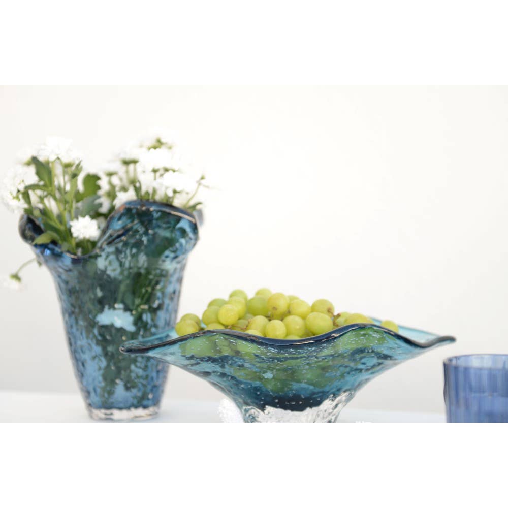 Grüne Weintrauben ruhen in der AULICA - Dekorative blaue Glasschale 33x18x10,5cm von AULICA, die neben einer blauen Glasvase mit weißen Blumen auf einer weißen Fläche steht; eine dekorative Schale und ein blauer Becher sind teilweise rechts zu sehen.