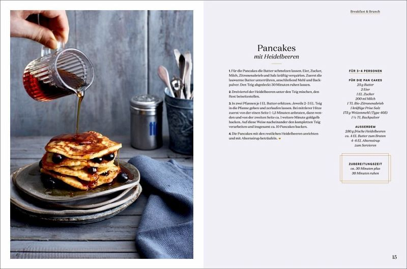 Ein Stapel Blaubeerpfannkuchen mit frischen Beeren und Sirup ist auf einem Teller abgebildet. Geschirr in einem Metallbecher und eine blaue Serviette vervollständigen die Szene - ideal für Fans von London Lovestory aus dem Hölker Verlag. Der Rezepttext erscheint auf der rechten Seite.