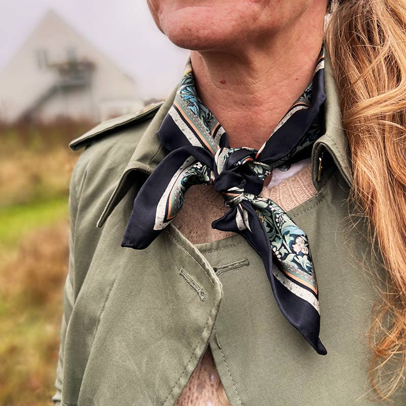 Eine Frau trägt einen olivgrünen Trenchcoat und den Koustrupco | Seidenschal Morris Bird 50 x 50 cm von Koustrupco, inspiriert vom William-Morris-Vogelmotiv, draußen mit unscharfem Gras und Gebäude im Hintergrund.