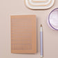 Das Tinne Mia Notizbuch A6 "Nougat Grid" mit FSC-zertifiziertem Papier, einer weiß-goldenen ovalen Ablage, einem lila Stift und einem transparenten lila Becher sind ordentlich auf einer hellbeigen Fläche arrangiert.