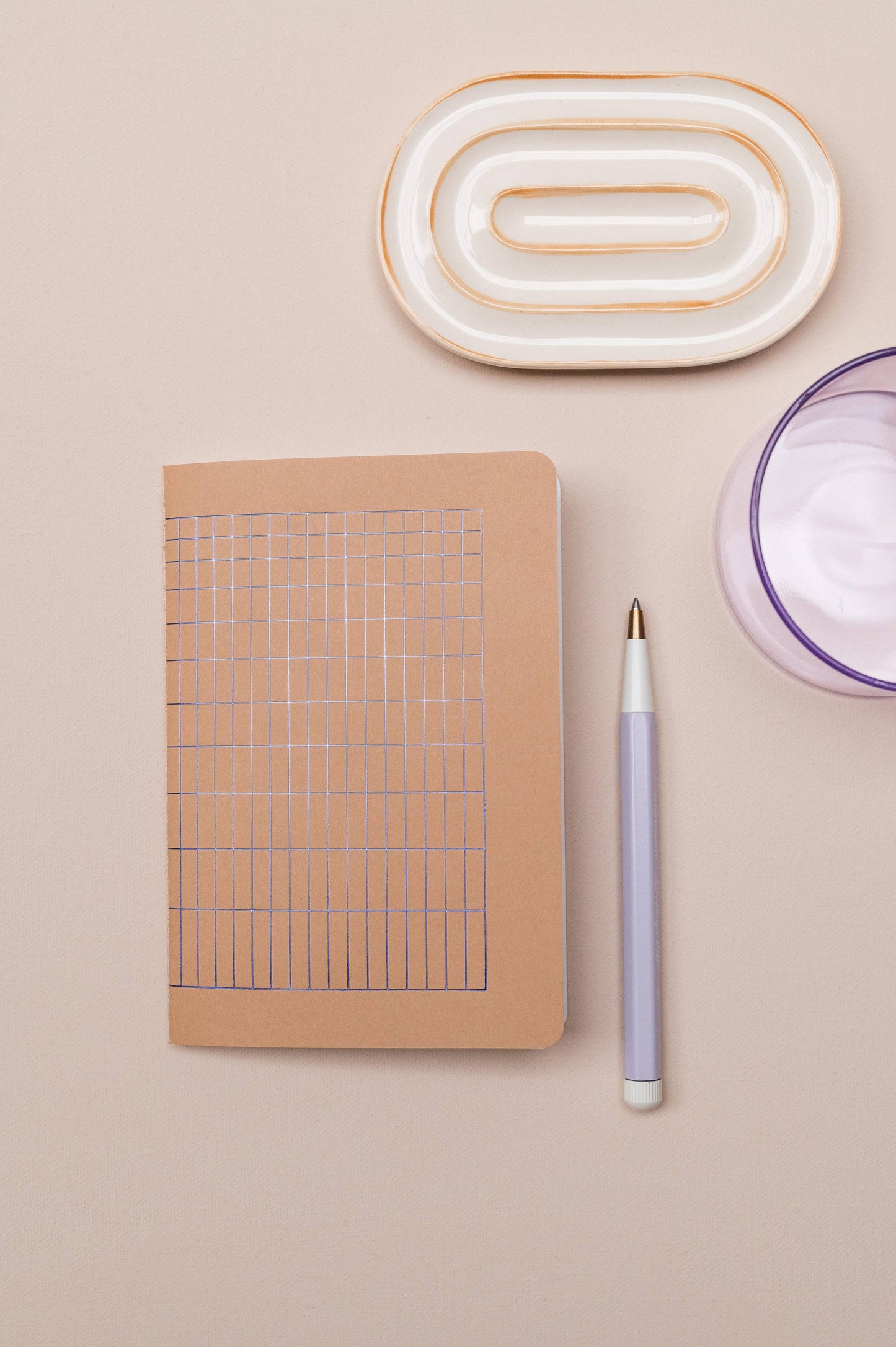 Das Tinne Mia Notizbuch A6 "Nougat Grid" mit FSC-zertifiziertem Papier, einer weiß-goldenen ovalen Ablage, einem lila Stift und einem transparenten lila Becher sind ordentlich auf einer hellbeigen Fläche arrangiert.