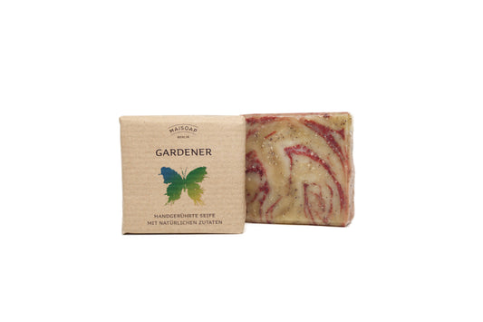 Ein Riegel maisoap berlin Gardener Seife mit roten und beigen Strudeln steht neben seiner braunen Papierverpackung mit der Aufschrift "GARDENER", die ein grün-gelber Schmetterling ziert - ein perfekter Leckerbissen für Gartenhände mit einer minzigen Duftnote.
