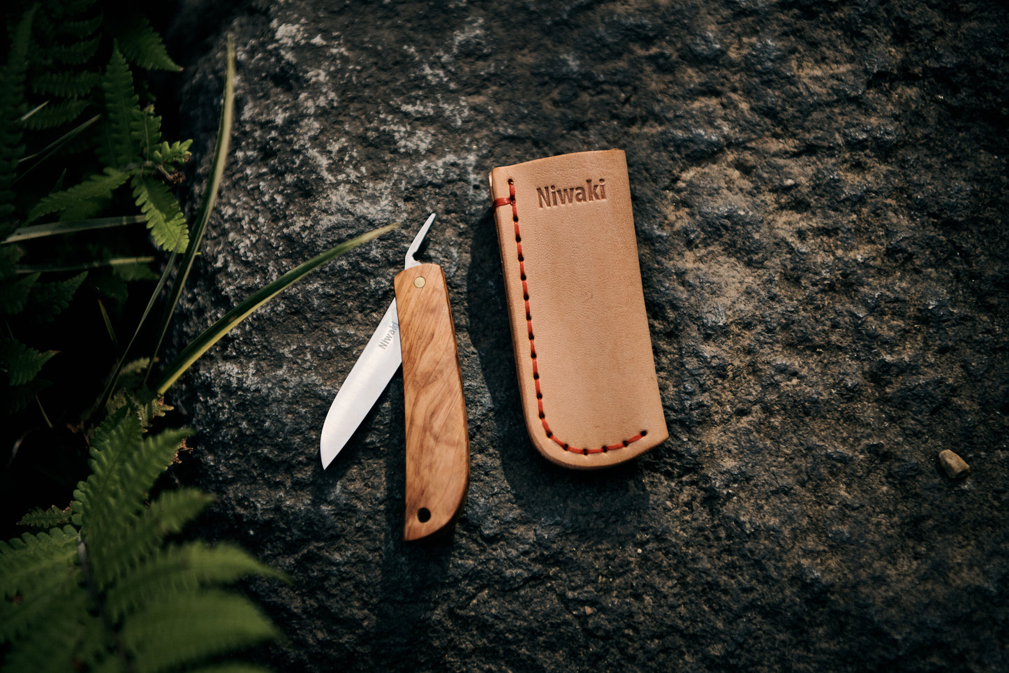 Ein Kotoh X Niwaki-Messer von Niwaki mit Holzgriff und teilweise geöffneter Klinge ruht auf einem Felsen neben einer Lederscheide mit roten Nähten und dem Niwaki-Logo. Grüne Farne rahmen die Szene ein und betonen den Bezug zum Gartenbau.