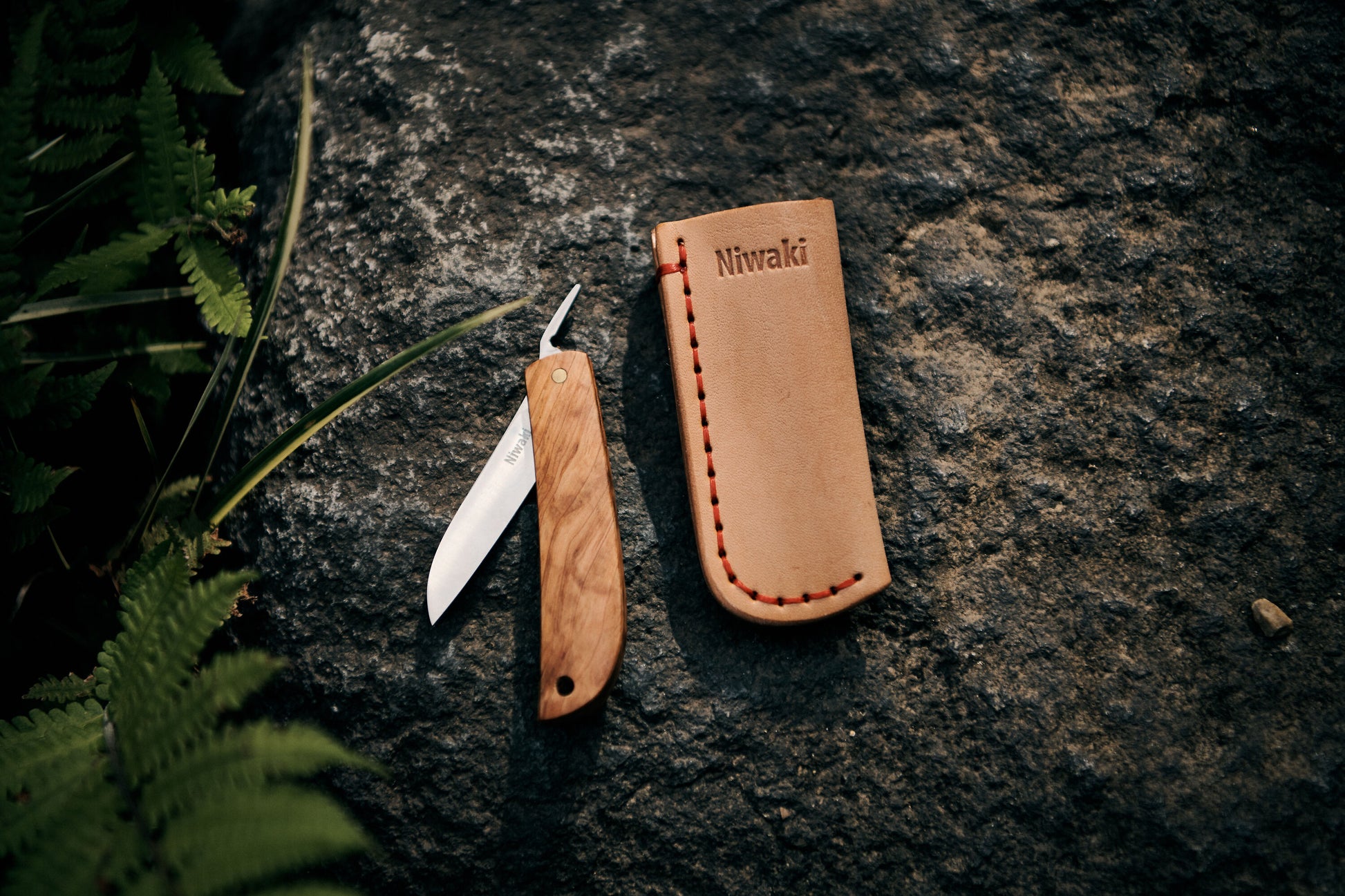 Ein Kotoh X Niwaki-Messer von Niwaki mit Holzgriff und teilweise geöffneter Klinge ruht auf einem Felsen neben einer Lederscheide mit roten Nähten und dem Niwaki-Logo. Grüne Farne rahmen die Szene ein und betonen den Bezug zum Gartenbau.