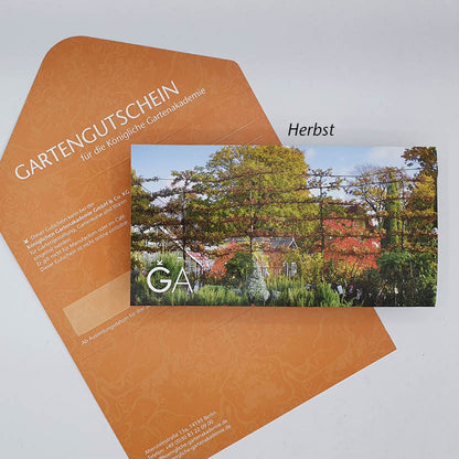 Ein orangefarbener Umschlag mit der Aufschrift „Gutschein für die Königliche Gartenakademie“ der Königlichen Gartenakademie, darin ein Gutschein mit einer herbstlichen Gartenszene, bunten Bäumen und darüber das Wort „Herbst“.