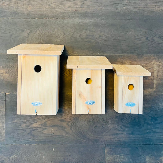 Drei Esschert Nistkästen aus zertifiziertem Erlenholz in verschiedenen Größen sind an einer dunklen Holzwand montiert, jeweils mit einem runden Eingang und einem blauen Aufkleber, perfekt für verschiedene Vogelarten.