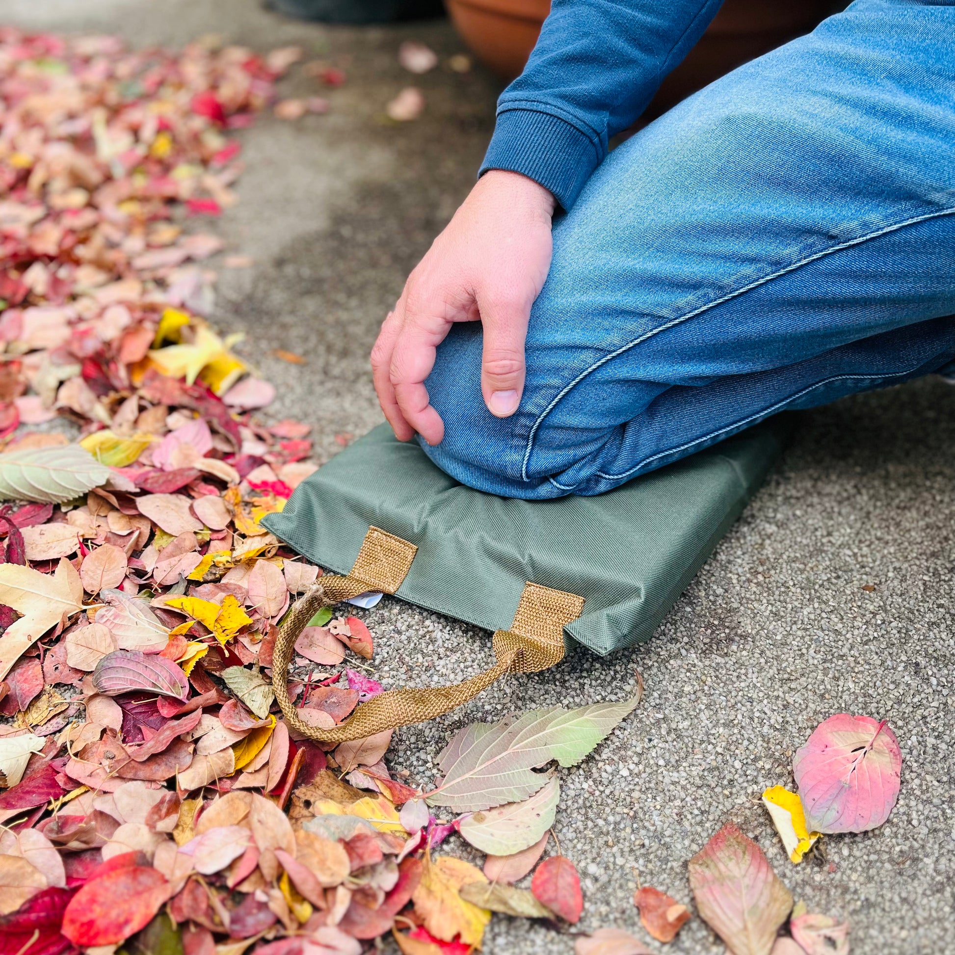 Eine Person kniet auf einem Esschert Kniekissen von Eschert Design mit Griffen für zusätzlichen Schutz bei der Gartenarbeit, neben einem Haufen Herbstlaub auf dem Pflaster. Sie trägt eine blaue Jeans und ein blaues langärmeliges Hemd.