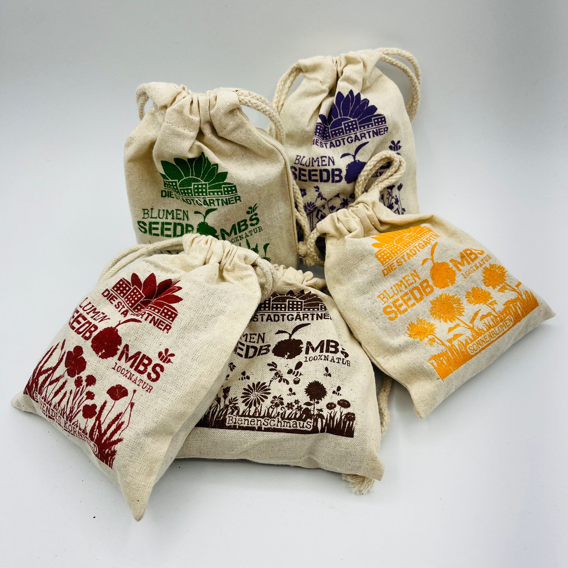 Vier kleine Stadtgärtner „Samenbomben – Wilde Wiese“ Kordelzugbeutel aus Naturstoff, jeweils mit Blumenaufschrift und farbenfrohen Wildblumenmotiven in Rot, Grün, Orange und Lila. Die Beutel stehen aufrecht auf weißem Untergrund.