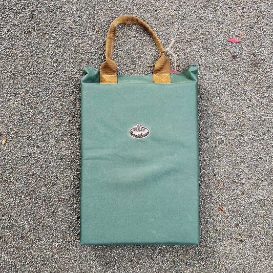 Das Esschert Kniekissen von Eschert Design, eine grüne, rechteckige Tasche mit braunen, geflochtenen Griffen und einem kleinen, ovalen Logo, ideal für die Gartenarbeit, ruht auf einer rauen, grauen, gekiesten Oberfläche.