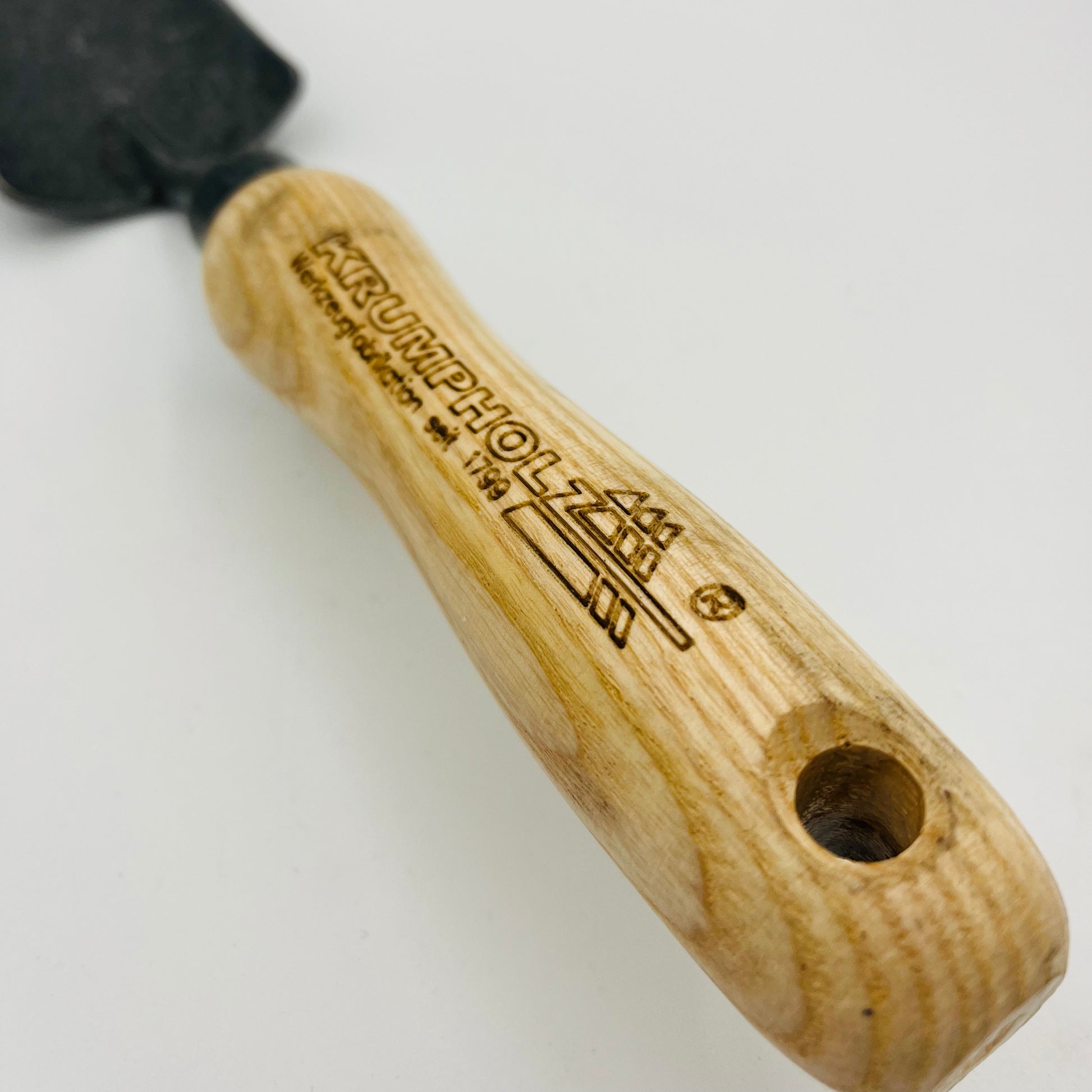 Eine Nahaufnahme der Krumpholz Pflanzkelle schmal zeigt den Holzgriff mit der Gravur „KRUMPHOLZ WERKZEUGE“ und dem Logo sowie einem Loch am Ende. Der Griff ist mit der schwarzen Metallklinge verbunden, die teilweise auf weißem Hintergrund sichtbar ist.