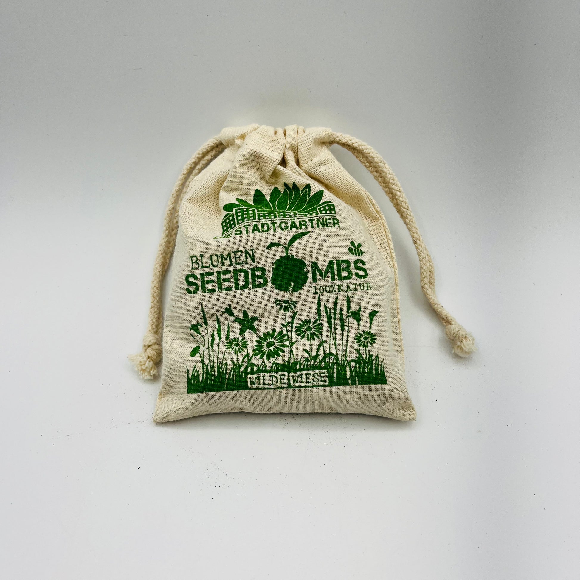 Ein kleiner Stoffbeutel mit Kordelzug von Stadtgärtner, verziert mit grünen Blumenillustrationen und deutschem Text. Mit der Aufschrift „Samenbomben - Wilde Wiese“ eignet er sich perfekt zum Aussäen von Wildblumensamen.