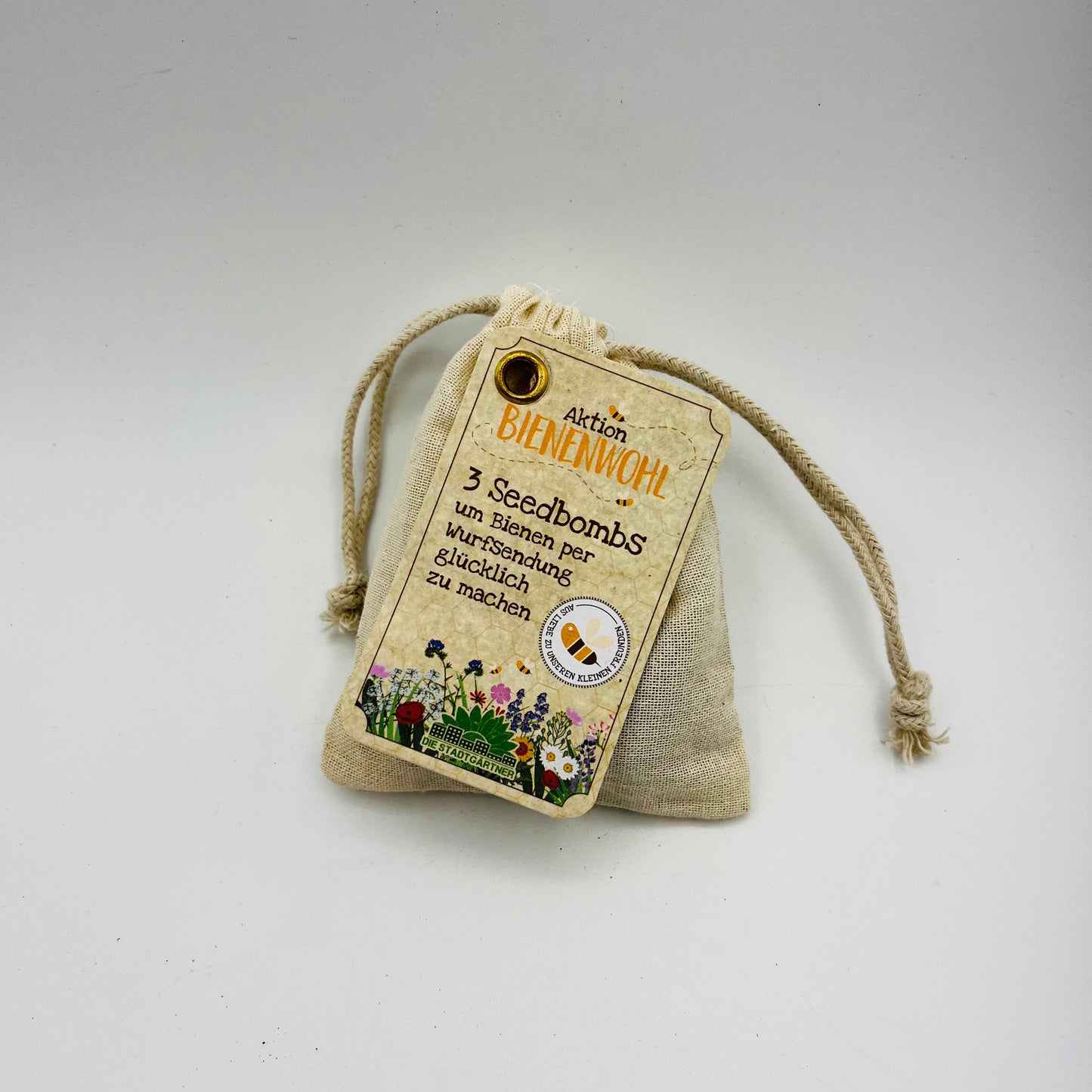 Ein kleiner beiger Kordelzugbeutel mit der Aufschrift „Stadtgärtner Bienenwohl Samenbomben“ enthält 3 bienenfreundliche Saatgut-Samenbomben, unten mit bunten Blumen und Bienen auf einem schlichten weißen Hintergrund dargestellt.