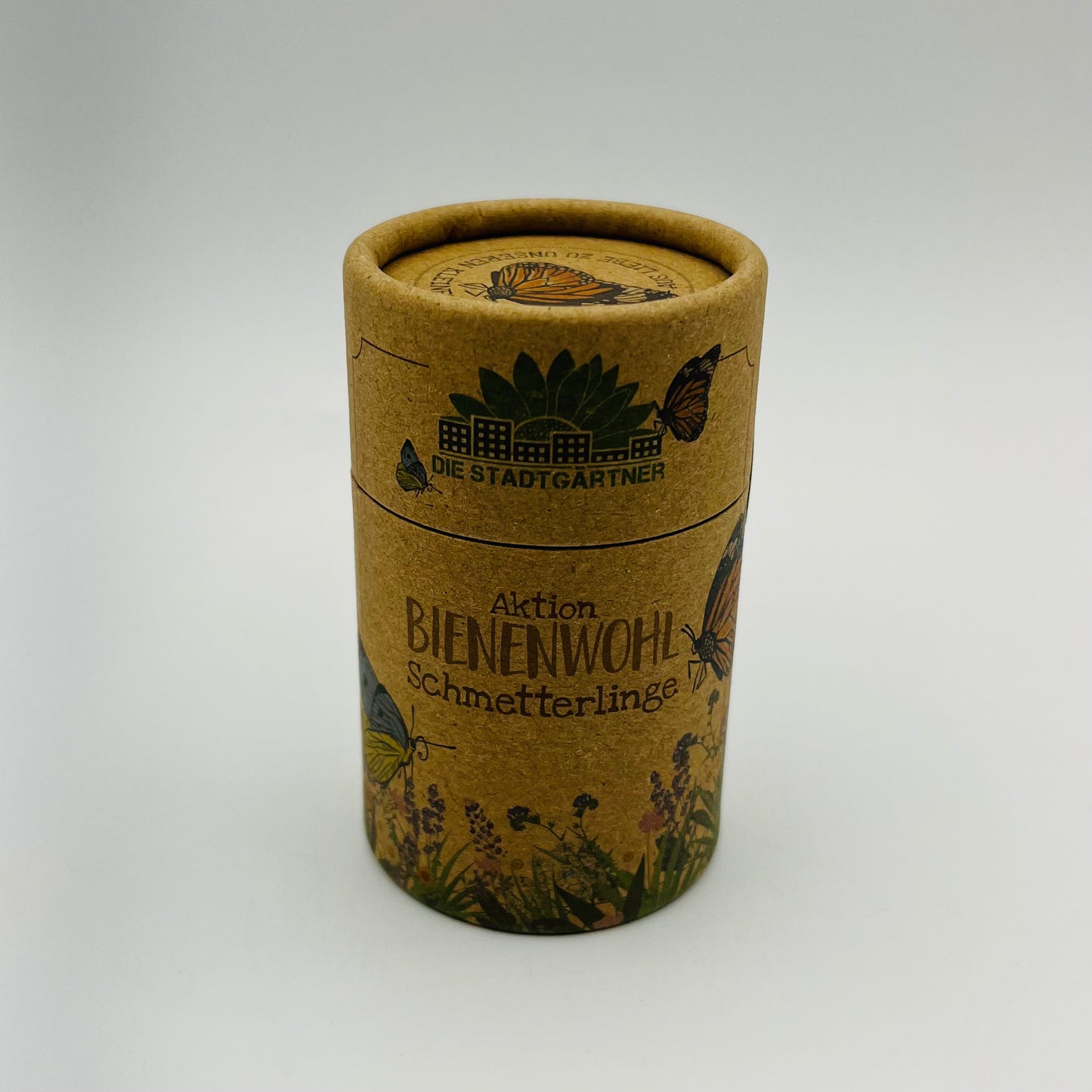 Ein zylindrischer Kartonbehälter von Stadtgärtner mit der Aufschrift „Bienenwohl-Saatgut – Schmetterlingswiese“, gefüllt mit Wildblumensamen und dekoriert mit Schmetterlingen, Bienen und Blumen auf hellbraunem Hintergrund.