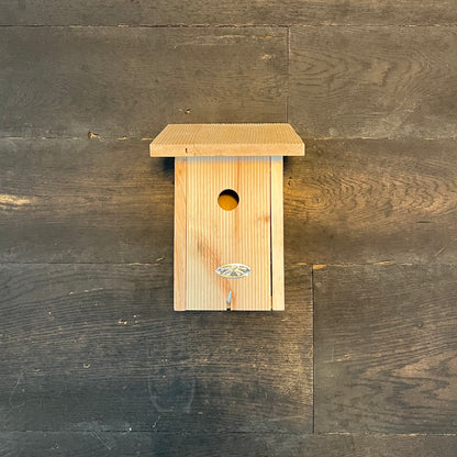 Der Esschert Nistkasten aus zertifiziertem Erlenholz verfügt über ein schräges Dach, einen runden Eingang und ein kleines dekoratives Emblem und ist somit ideal für verschiedene Vogelarten. Er wird an einer dunklen Holzwand montiert.