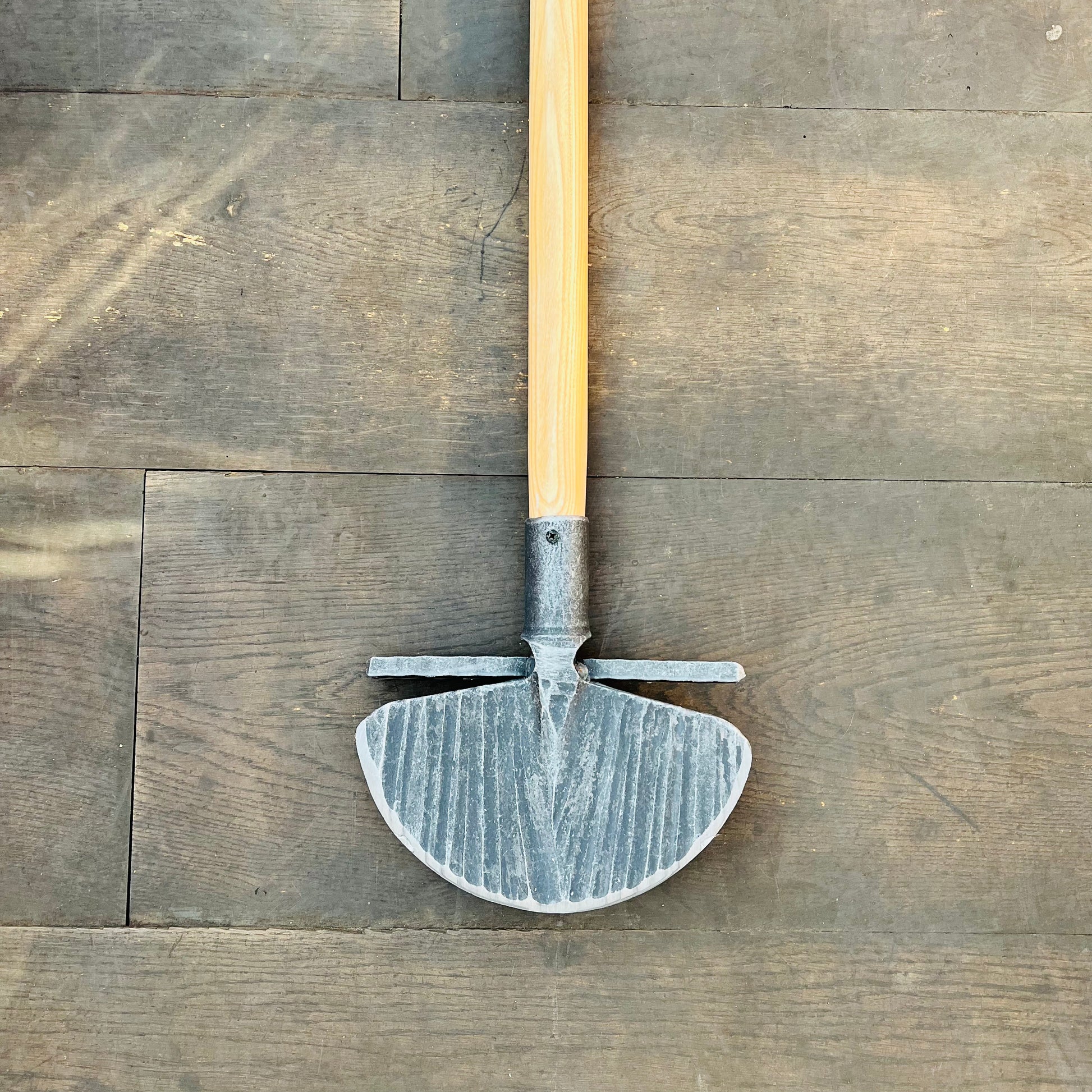 Ein Krumpholz Rasenkantenstecher 85cm mit einem Holzgriff und einer halbrunden, geriffelten Metallklinge mit zwei kurzen horizontalen Stäben liegt flach auf einem Holzboden.